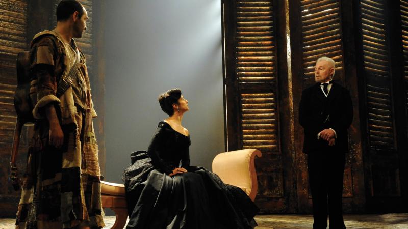Female actor in a black velvet dress, seated, looks defiantly at the male actor in a dark suit, during a scene of Twelfth Night