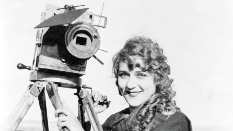 Pickford in a plaid coat, standing with one of her cameras and smiling at the camera