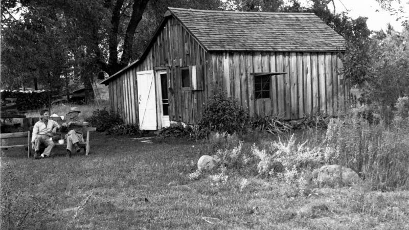 Black and white photo of two men on a bench outside a shack