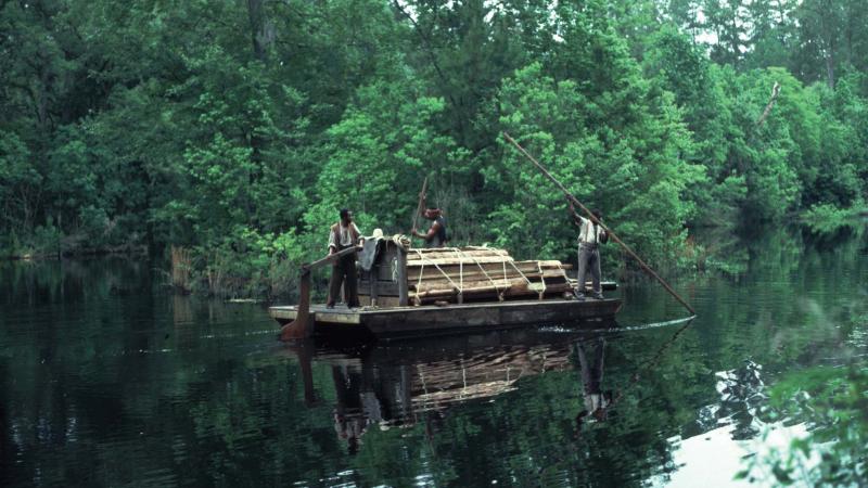 Northup steers a raft loaded with logs down a river surrounded by trees