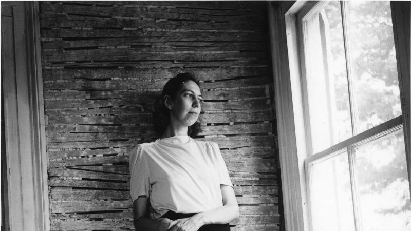 Welty in a white blouse and black skirt, leaning against a brick wall and looking out of a large window to her left