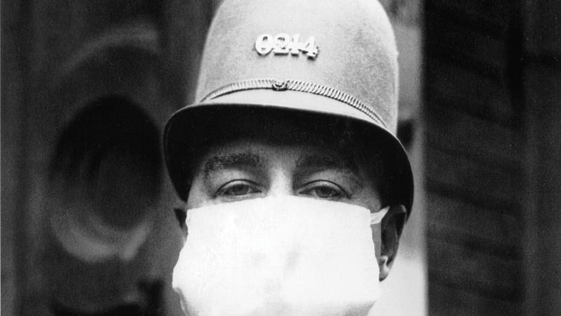 A police officer, in uniform and rounded helmet, has his entire face, other than the eyes, covered by a white cotton medical mask