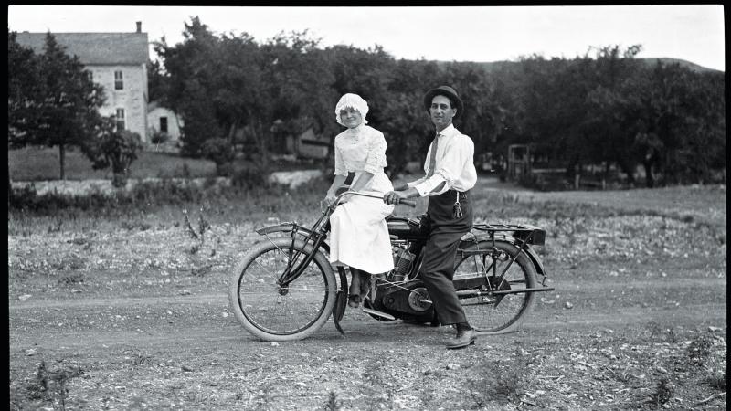 Kratzer, in a white shirt and black hat, sits on a motorcycle behind a woman in a white dress and cap, on a dirt road