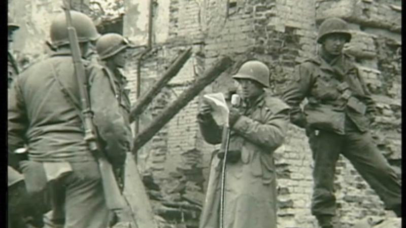 Three armed and uniformed soldiers watch Plambeck, also uniformed, speak into a microphone while reading off of a paper, in the ruins of a stone building