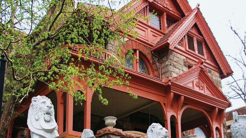 Color photo of an ornate, wide-brick home with red painted trim and columns. 