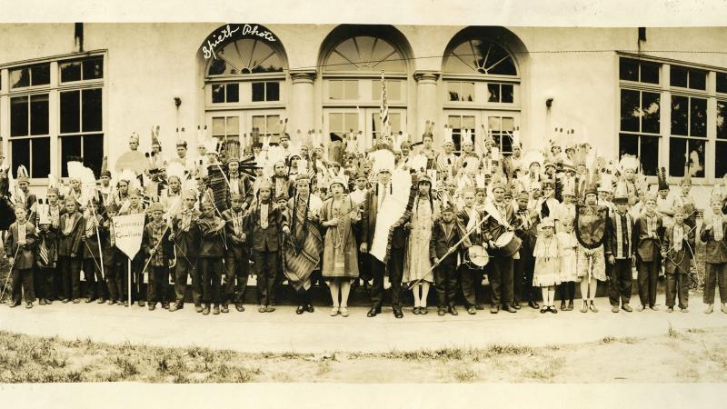 Sepia-colored photograph of a tribe of Native Americans standing in front of a large building.