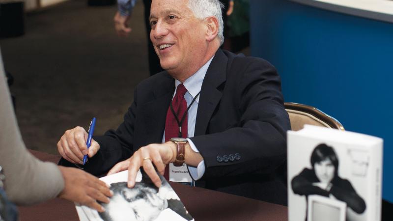 Color photo of Walter Isaacson at a book signing for his best-seller. He is seated at a desk and smiling at one of his visitors.