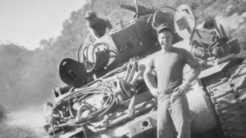 McInroe stands with his hands on his hips, pipe in his mouth, in front of his tank