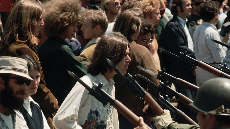 Color photo of protesters facing army bayonets at a checkpoint. 