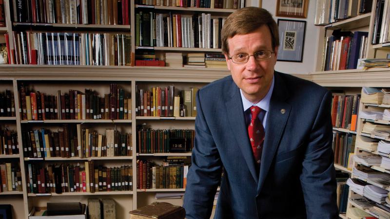 Photo of a man in a suit leaning over a rare book in a library, while looking at the camera.