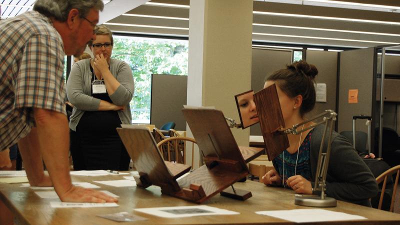 Photo of a woman seated at a desk, examining a rare book through an optical device. Two onlookers watch.