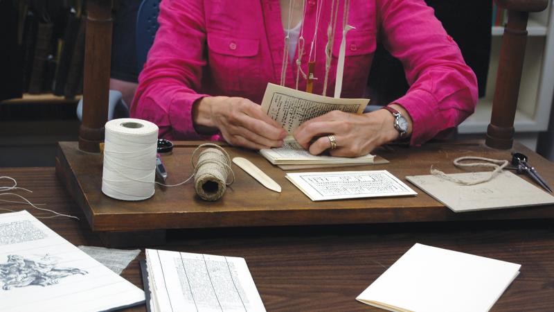 Photo of a woman wearing a pink blazer seated at a desk, binding a rare book.