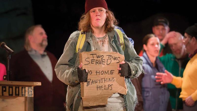 An actor holds up a sign reading "served two tours, no home, anything helps" during a scene