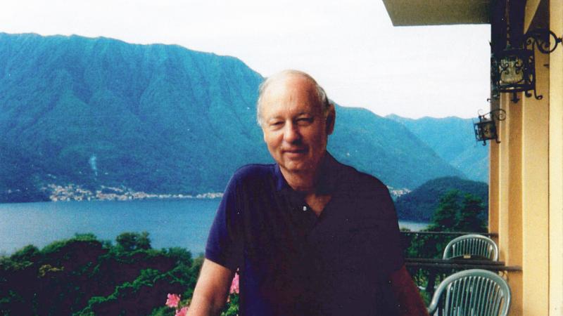 Color photo of Jules Witcover standing on the balcony of a house which overlooks a large bay and mountains in the background.