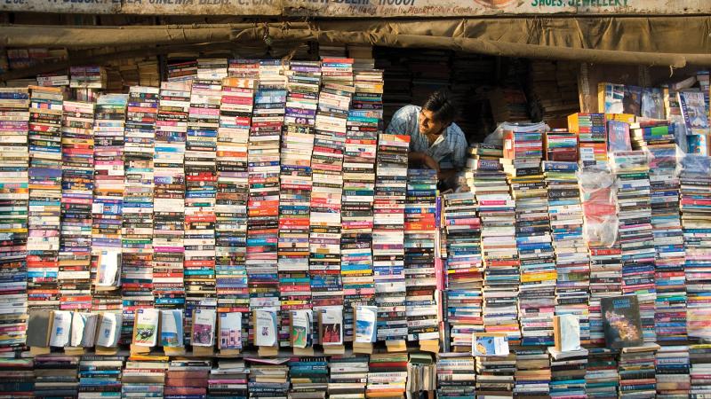 Color photo of a man in Delhi, India, peering out from behind a wall of books which comprises his storefront.