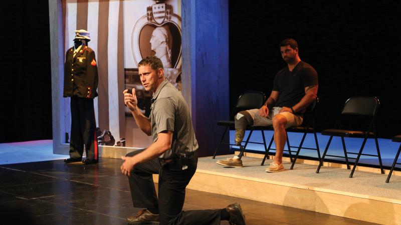 Color photo of two veterans, one kneeling while performing on stage, the other sitting in the background. 