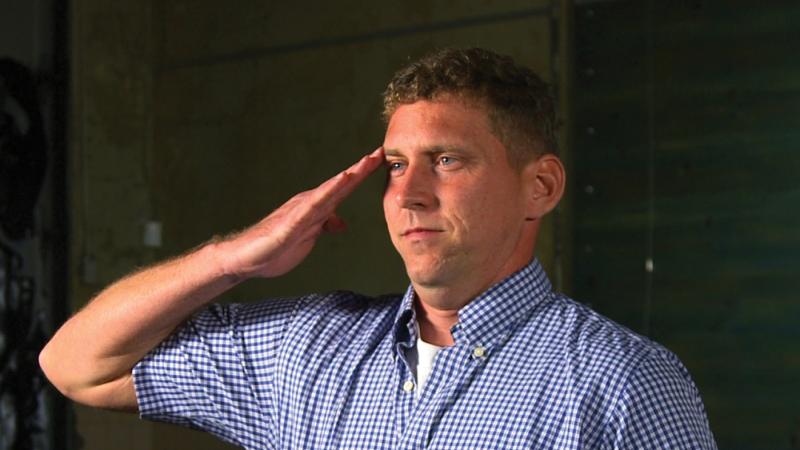 Color photo of a man wearing a blue shirt saluting an audience.