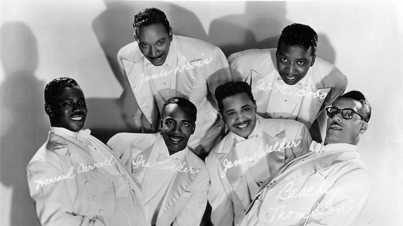 Black and white photo of a musical group wearing white tuxedos and posing for a photograph at irregular angles.