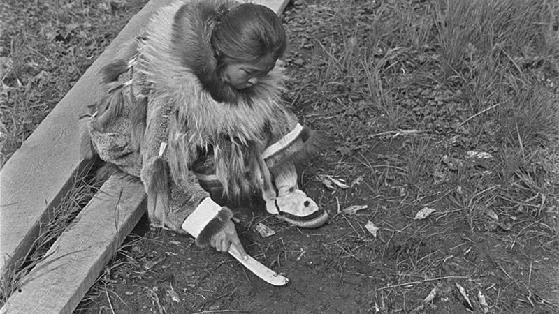 Black and white photo of an Indian woman sitting on the ground with a knife-shaped tool in her hand.