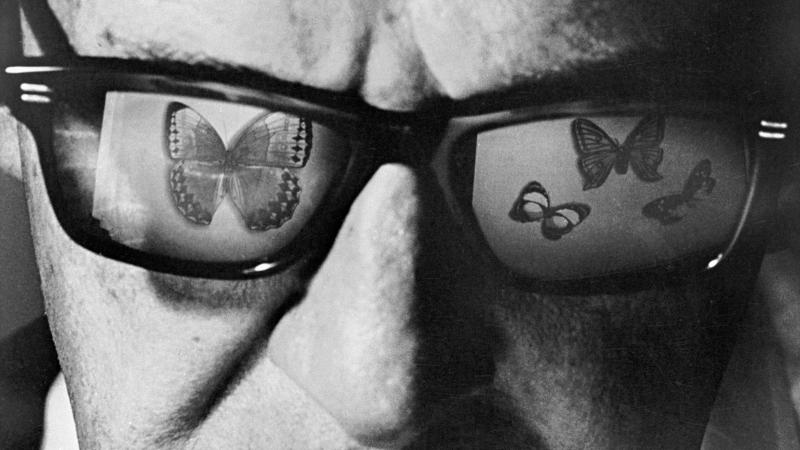 black and white close-up portrait of a man wearing glasses, which reflect butterflies