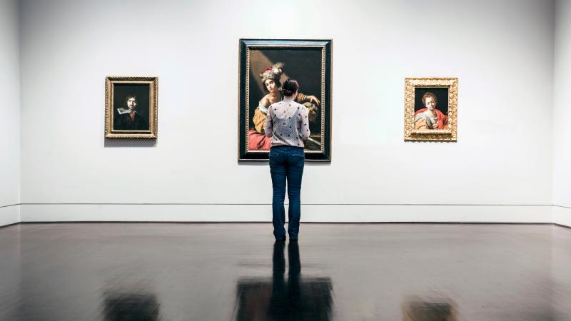 Photograph of woman standing in museum, three paintings on the wall