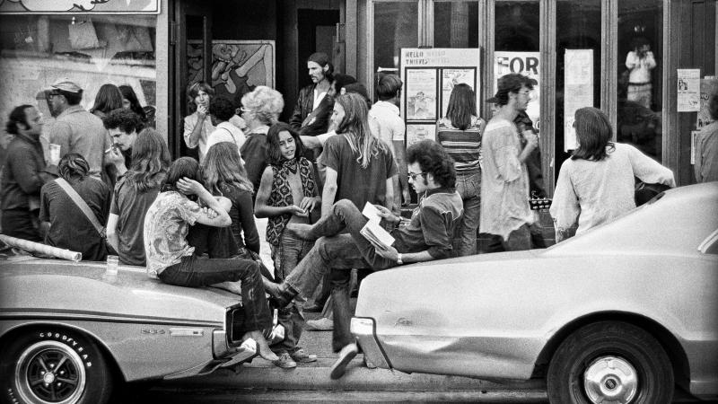 Black and white photograph of people standing around and lounging on cars