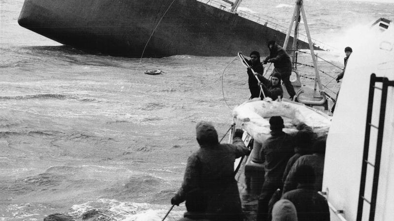Black and white photo of men pulling in a life raft from a sinking ship.
