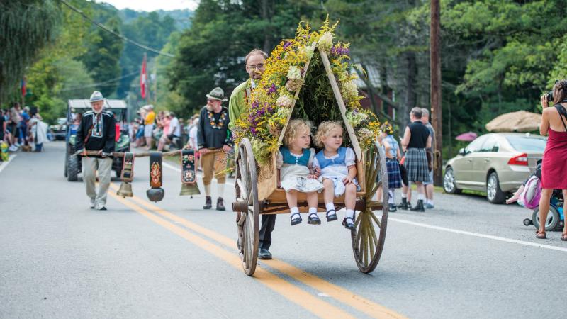 Color photo of a street festival with a man pushing a decorated carriage with two children on board.