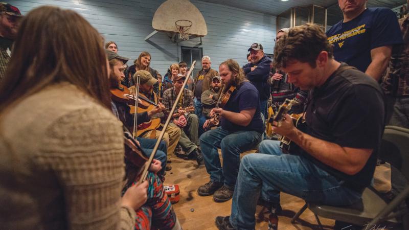 Color photo of a group of musicians sitting in chairs in a circle, playing a variety of instruments including guitars and violins.