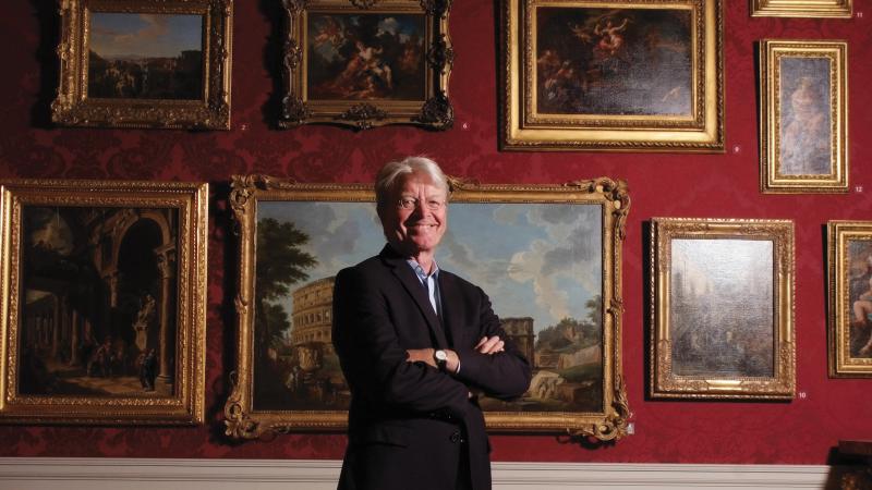 Color photo of Gary Vikan standing before a wall of paintings. He wears a black blazer and khaki dress pants.