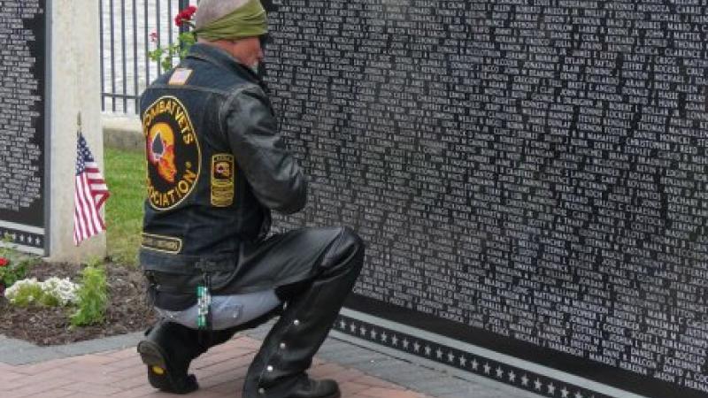 A man in jeans and a motorcycle jacket kneels before the Vietnam War Memorial.