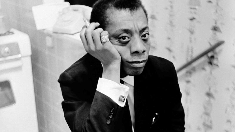 Black and white photo of James Baldwin, sitting backwards on a foldable chair, in a kitchen. He's is wearing a black suit with a white shirt and cufflinks. He is holding a cigarette in his right hand with a bold pink ring.