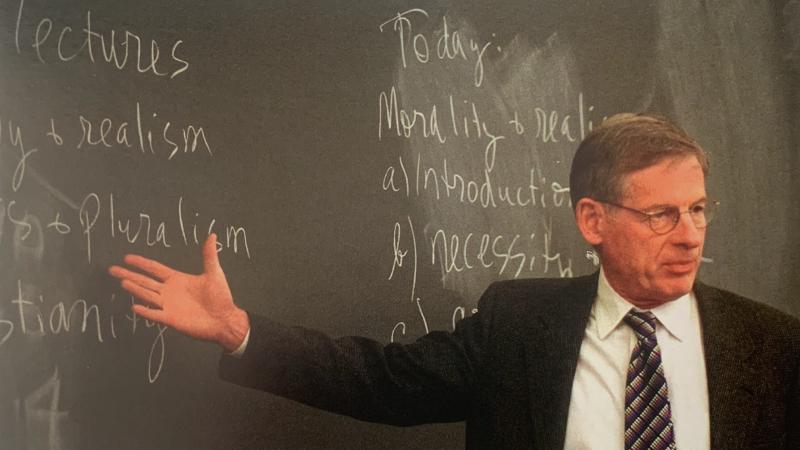 Mansfield lectures to a class at Harvard University. He is pointing at a green chalkboard in a black suit, patterned tie, white shirt, and glasses. He is facing the class.