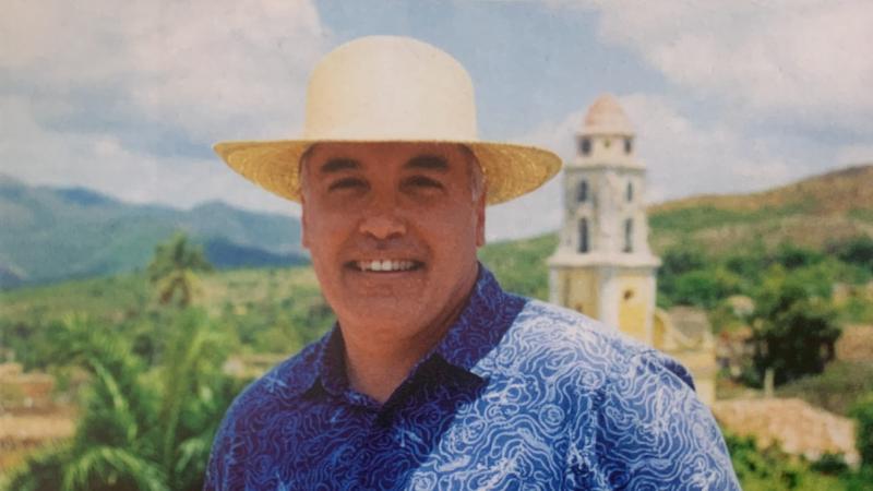 Portrait of David Tebaldi in a blue, decorative shirt and straw hat., with a countryside background.
