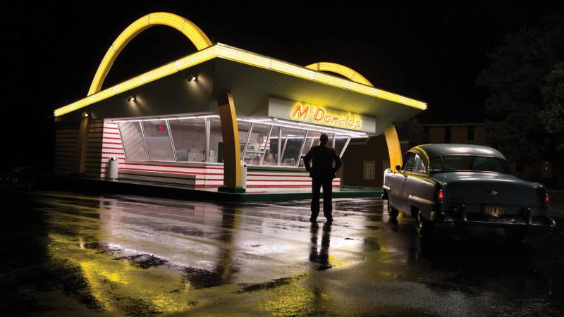 a movie still shows Ray Kroc in front of a nighttime golden arches.