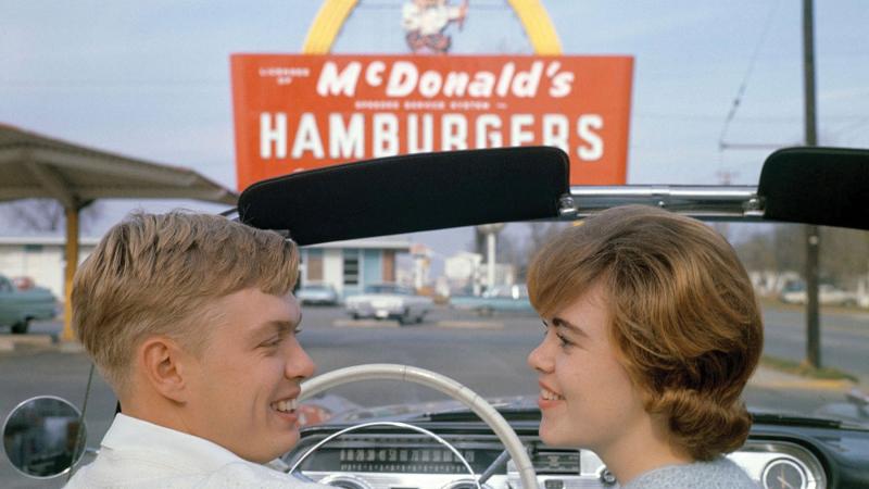 two white teenager sit in a car in front of a 1950s McDonald's sign.