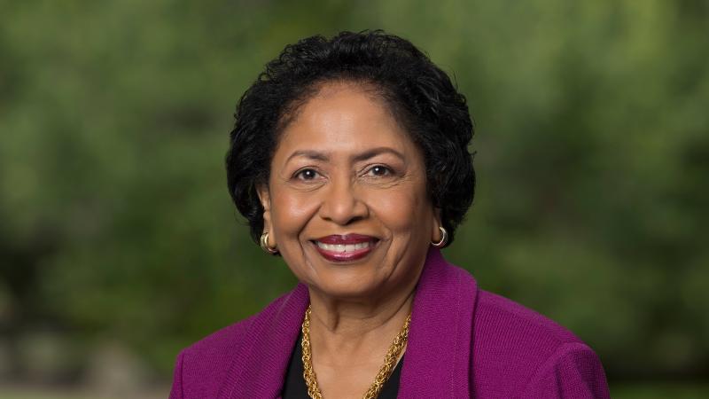 Dr. Ruth J. Simmons in a purple jacket and gold chain against a dark, leafy background