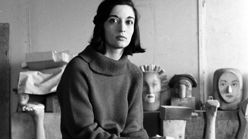 Marisol Escobar seated on stool with pieces of artwork behind her. 